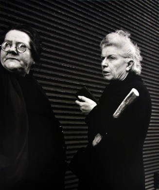 A black and white photograph of two older women standing by a horizontally ribbed wall.