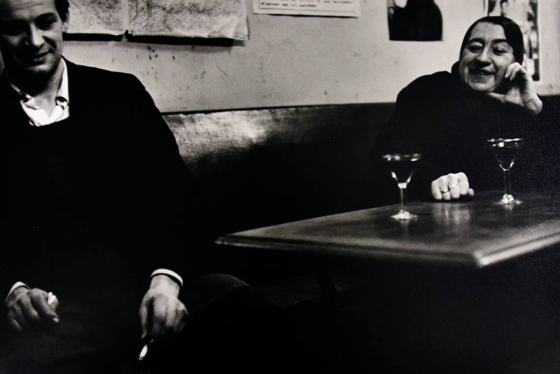 A black and white photograph of a man sitting adjacent to a woman by two sherry glasses on the …