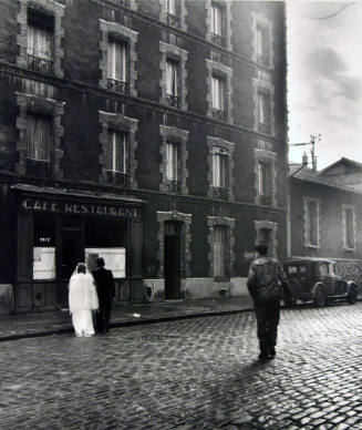 A black and white photograph of a man walking in the middle of a street toward newlyweds in fro…