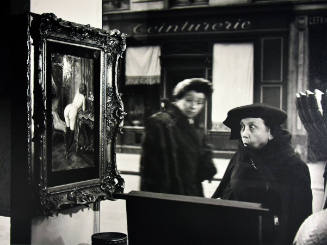 A black and white photograph of two women peering into a store window containing a painting of …