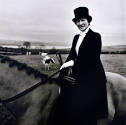 A black and white photograph of a woman in a long black dress and black top hat sitting side-sa…