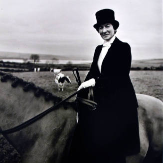 A black and white photograph of a woman in a long black dress and black top hat sitting side-sa…