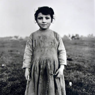 A black and white photograph of a girl with dark hair and a long sleeved, dirty dress standing …