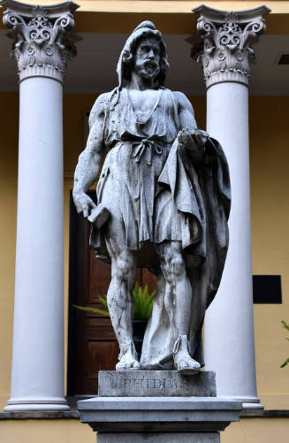 A long-haired male wearing a tunic tied at the waist, holding a cloak over his proper left arm …