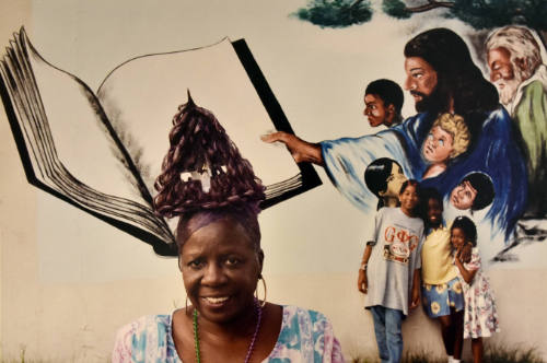 A woman with a triangular coiffure stands in front of a mural of Jesus Christ holding out a boo…