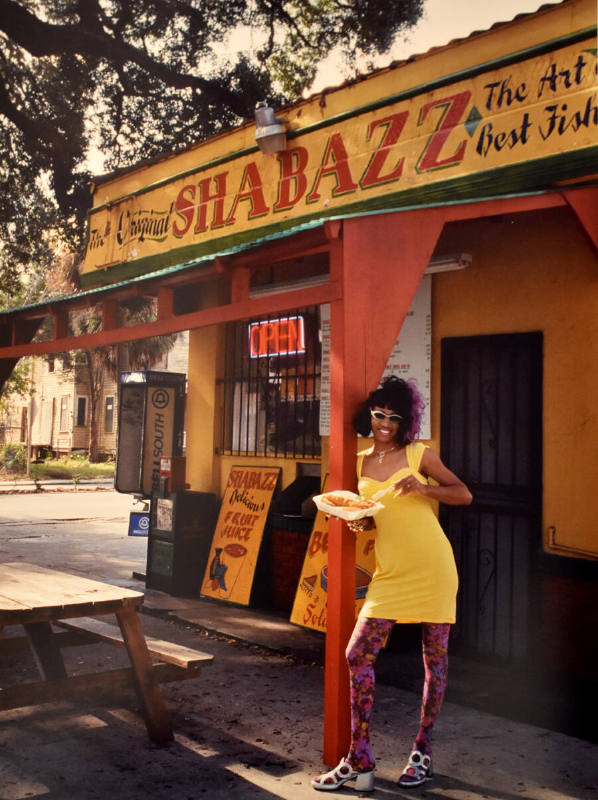 A woman in a bright yellow dress standing in front of a yellow building with green trim and red…