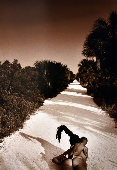 A nude woman with a tall column of hair lying in the middle of a dirt road with an infant. 