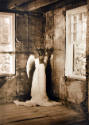 A woman in a long white gown with wings stands staring into a corner of an old sparse room.