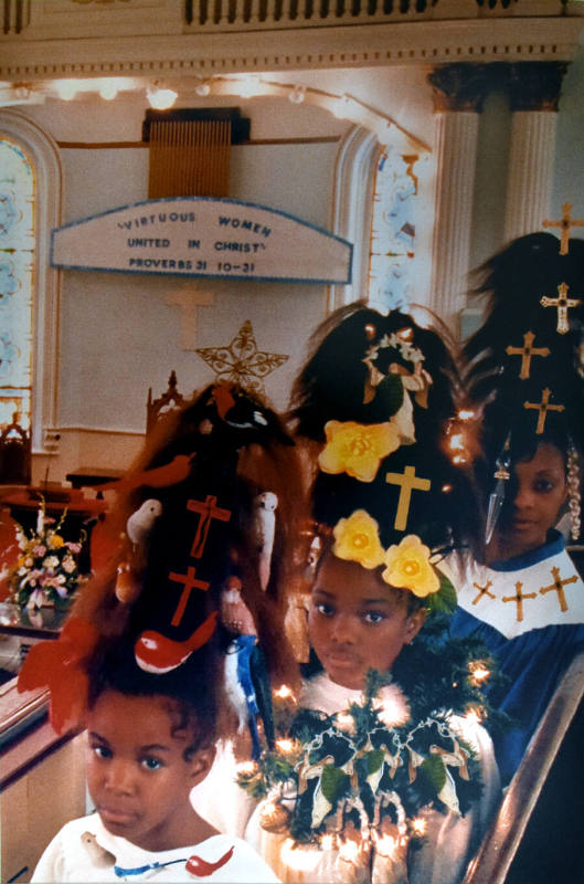 Three girls wearing choir robes with large triangular hairdos covered in crosses, birds, and fl…