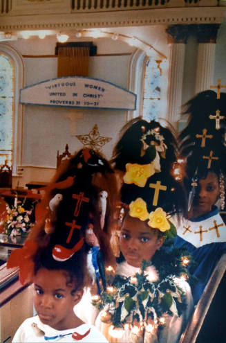 Three girls wearing choir robes with large triangular hairdos covered in crosses, birds, and fl…