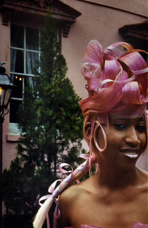 A woman with pink curled hair and a blonde braid standing in front of a pink-toned building.