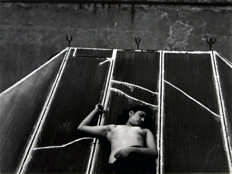 A black and white photograph of a topless female laying on her back on a roof-like structure.