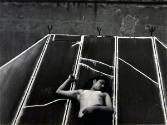 A black and white photograph of a topless female laying on her back on a roof-like structure.