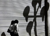 A black and white photograph of two women walking passed a corrugated wall with a large cast sh…