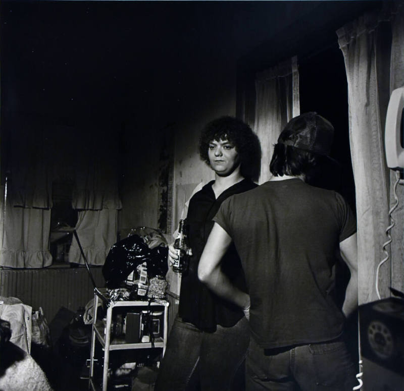 A black and white photograph of a man and woman in a room. The man is looking out a window.