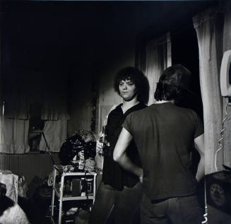 A black and white photograph of a man and woman in a room. The man is looking out a window.
