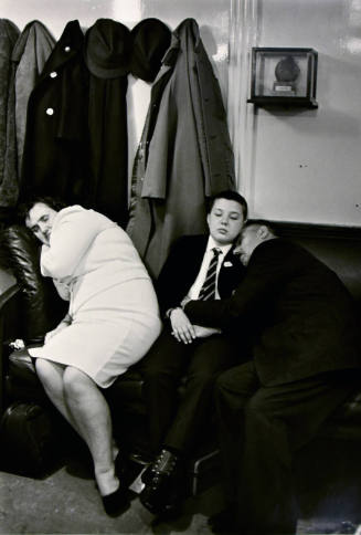 A black and white photograph of three figures sleeping on a sofa by a coat rack.