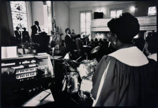 A black and white photograph of a woman in choir robes behind an organ and drum set looking out…