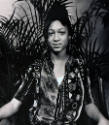 A black and white photograph of a seated young woman in a patterned shirt in front of a palm tr…