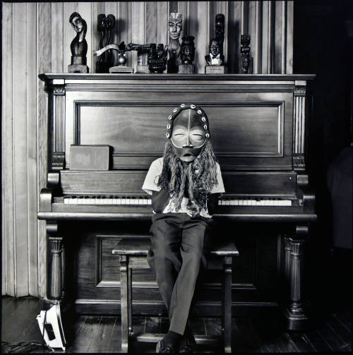 A black and white photograph of a little boy seated on a piano bench in front of a piano holdin…
