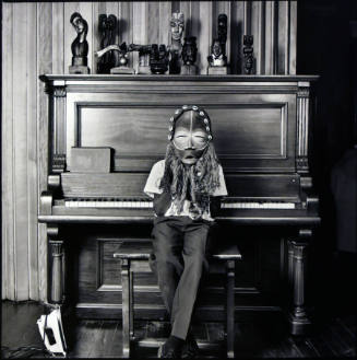 A black and white photograph of a little boy seated on a piano bench in front of a piano holdin…