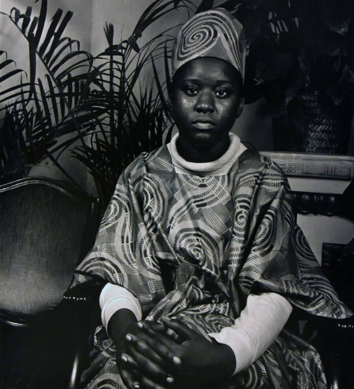 A black and white photograph of a seated young woman in a swirl-patterned garment with her hand…