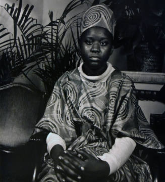 A black and white photograph of a seated young woman in a swirl-patterned garment with her hand…