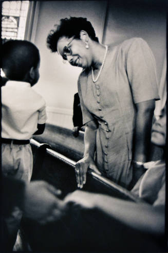 A black and white photograph of the back of a little boy standing on a church pew looking at a …