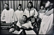 A black and white photograph of young women in white choir robes singing with two holding saxop…