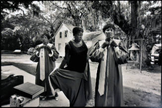 A black and white photograph of three women donning their choir robes by a tree-lined road with…