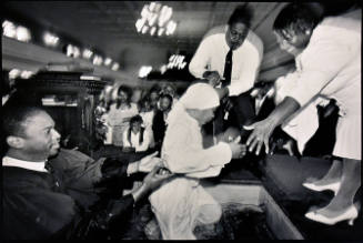 A black and white photograph of a young woman being helped out of a baptismal font or pool by t…