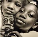 A black and white photograph of two girls holding a carved figure by their faces.