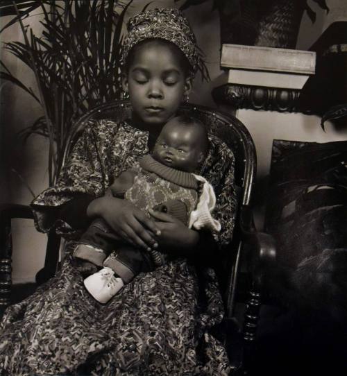 A black and white photograph of a seated girl in a patterned garment holding a plastic doll.