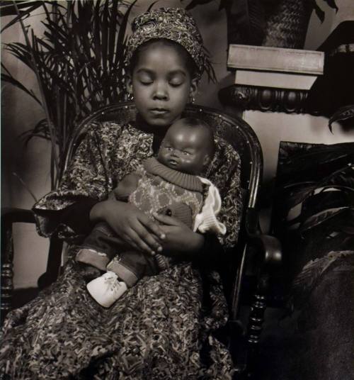 A black and white photograph of a seated girl in a patterned garment holding a plastic doll.