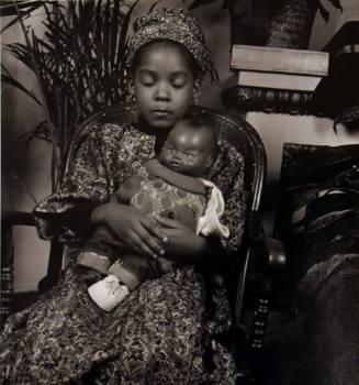 A black and white photograph of a seated girl in a patterned garment holding a plastic doll.