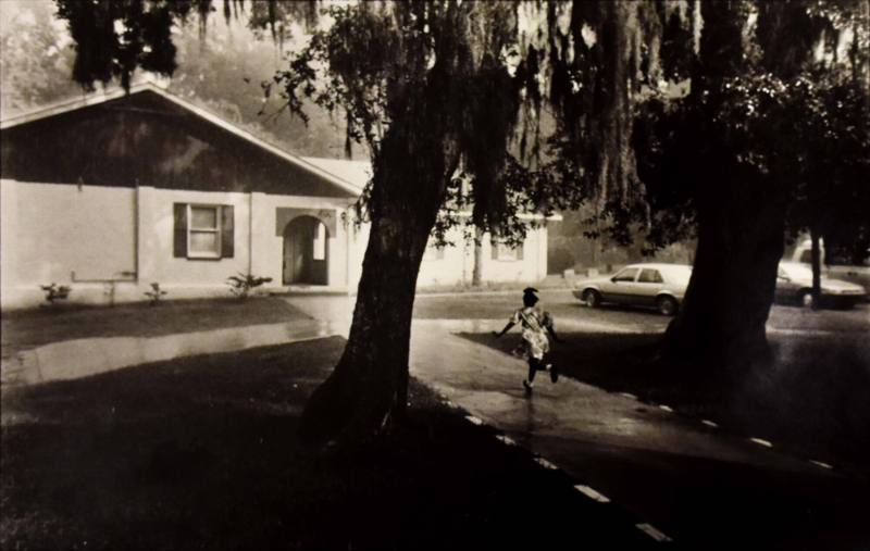 A black and white photograph of a girl running down a sidewalk between two trees towards a smal…