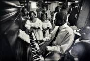 A black and white photograph of a man playing the piano next to three girls in white choir robe…