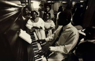A black and white photograph of a man playing the piano next to three girls in white choir robe…