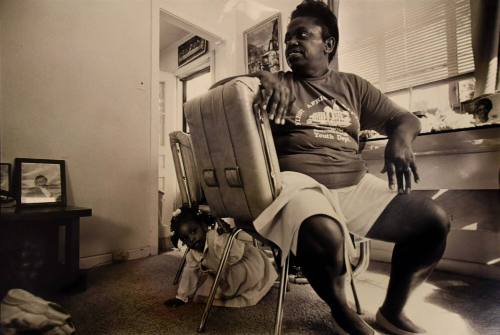 A black and white photograph of a woman seated on a chair with her head facing the left while a…