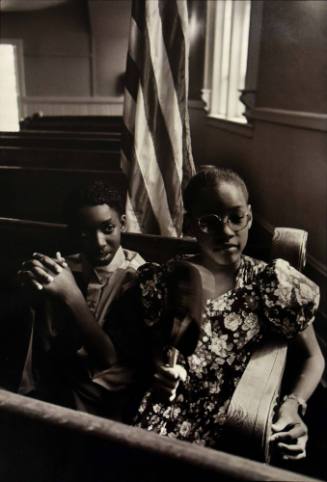 A black and white photograph of a boy and a girl seated in a pew in front of an American flag.