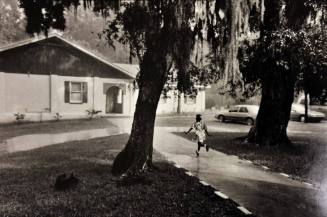 A black and white photograph of a girl running down a sidewalk between two trees towards a smal…