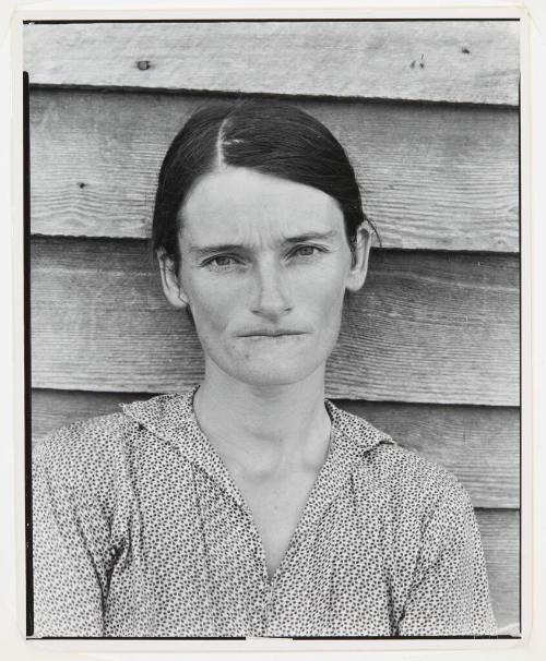 A black and white bust-length portrait of a woman in front of a clapboard house.