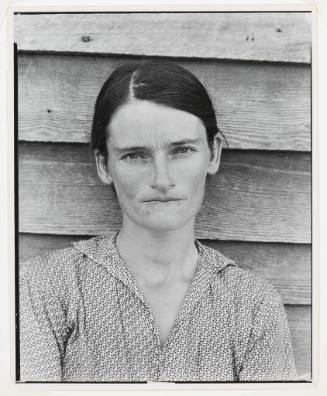 A black and white bust-length portrait of a woman in front of a clapboard house.