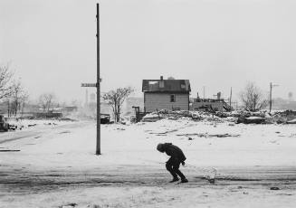 A black and white winter scene in a residential area with an industrial background. The figure …