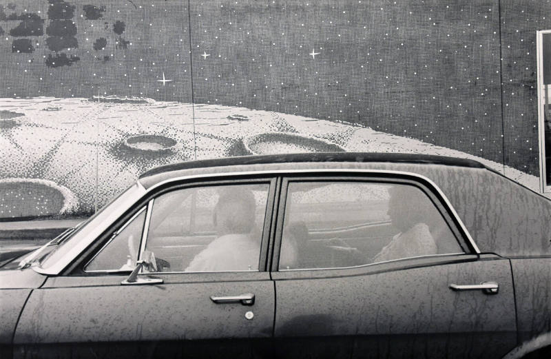 A black and white photograph of a man sitting in the driver's seat of a car, his head turned aw…