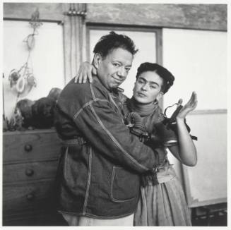 Looking towards the camera, Diego Rivera embraces Frida Kahlo while she holds a gas mask in her…