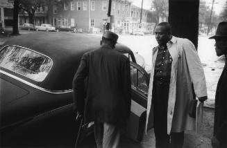 A black and white photograph of a man in a trench coat holding a car door open for a man in a d…