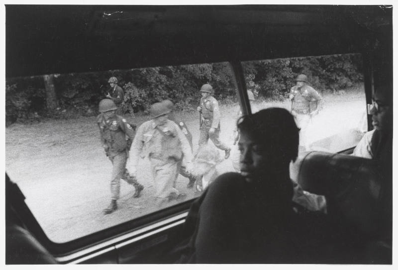 A black and white photograph of Freedom Riders in a bus looking out at soldiers walking along t…