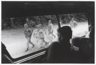 A black and white photograph of Freedom Riders in a bus looking out at soldiers walking along t…