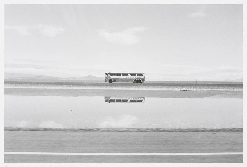A greyhound bus travels across a landscape and is reflected in a body of water.